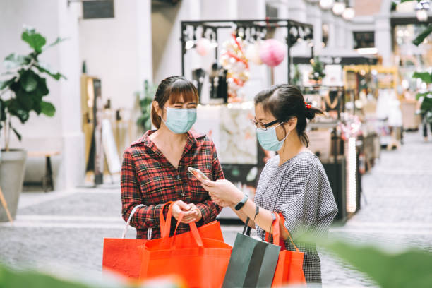 2人のアジアの女性がスマートフォンを見てショッピングストアを検索する保護フェイスマスクを着用 - ストック写真 - ミディアムショット ストックフォトと画像