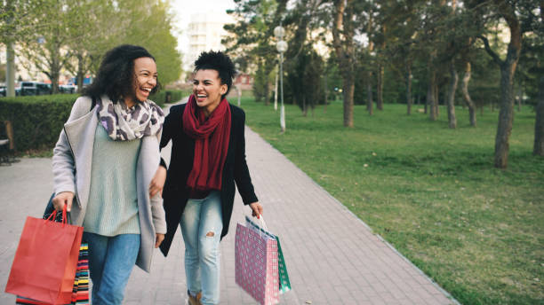 two attractive women dancing and have fun while walking down the park with shopping bags. Happy young friends walk after visiting mall sale stock photo