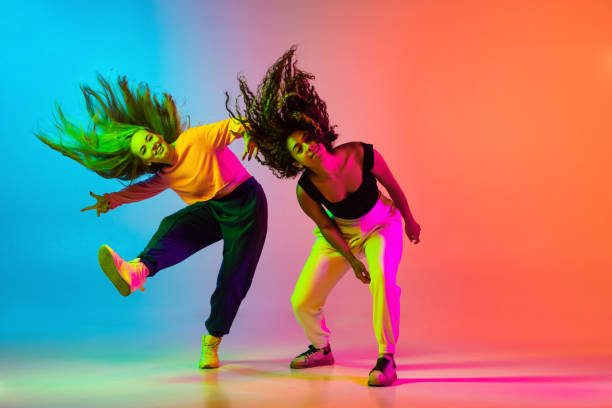 Two beautiful hip-hop girls dancing on gradient blue orange backlground in neon Dynamic lifestyle. Two beautiful hip-hop dancers in motion on gradient blue orange backlground in neon. Sport achievement, expression. Concept of dance, youth, hobby, dynamics, movement, action, ad fluorescent light photos stock pictures, royalty-free photos & images