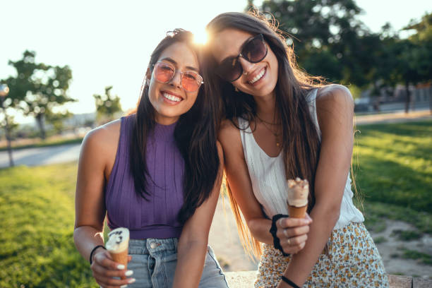 Two beautiful young women eating ice cream while having fun walking through the park in the city. stock photo