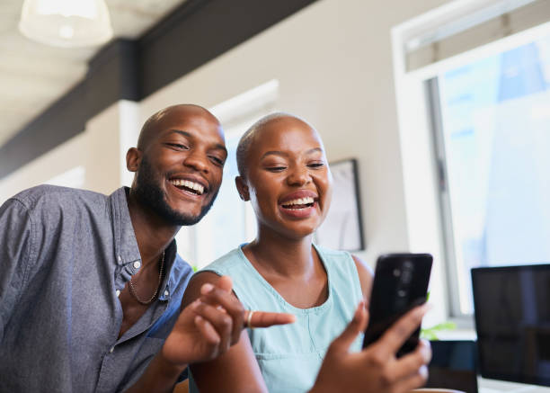 Two Black colleagues watch a funny video on cellphone point and laugh stock photo