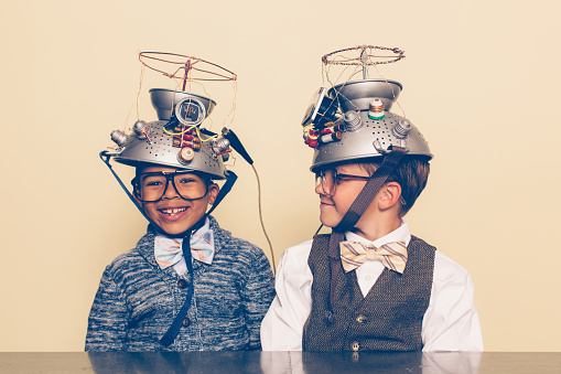Two nerd boys dressed in casual clothing, glasses and bow ties experiment with a homemade science project. They are both smiling and sitting at a table, and one is looking at the other with helmets on their heads in front of a beige background. Retro styling.