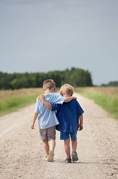 Two Boys Walking Down a Gravel Road Two young boys with their arms around each other walking down the country road. Vertical colour image. Copyspace. Back view of two cute Caucasian boys. Additional themes include friendship, bonding, talking, care, love, siblings, brothers, walking, rural, exercising, togetherness, cute, humour, summer, relationships, and affection.  arm around stock pictures, royalty-free photos & images