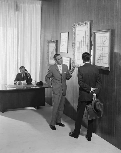 Two businessman discussing at bar chart while another man using telephone in background  1964 stock pictures, royalty-free photos & images