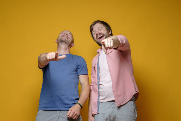 Two Caucasian men in casual clothes laugh mercilessly at someone and point with their index finger. Yellow background. stock photo