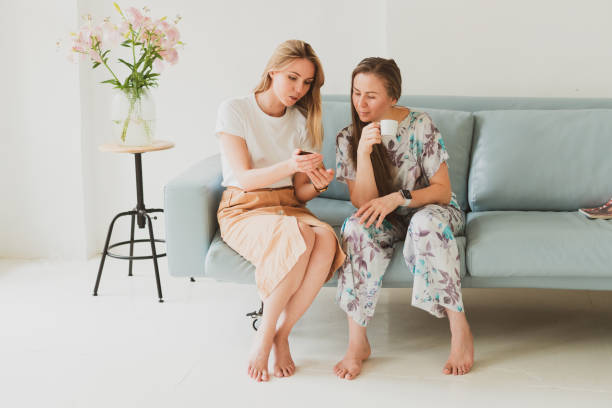 two charming young women chatting at home on the couch, drinking coffee and looking for information on the phone stock photo
