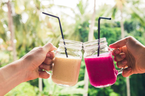 Two colorful fruit shakes in hands. Summer and tropical mood. Cold blended drinks, banana and dragon fruit smoothie. Clink glasses by couple hands stock photo