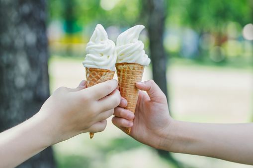 Two cones with creamy ice cream in children's hands. Children walk in the park in summer and eat sweets. Horizontal photo with place for text.