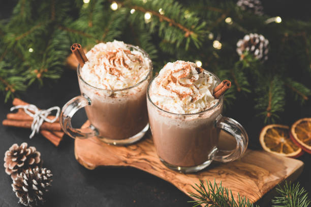 Two cups of hot chocolate with whipped cream and cinnamon Two cups of hot chocolate with whipped cream and cinnamon on wooden serving board surrounded with fir tree, Christmas lights, cinnamon and pine cones. Cozy Christmas Drink comfort food stock pictures, royalty-free photos & images