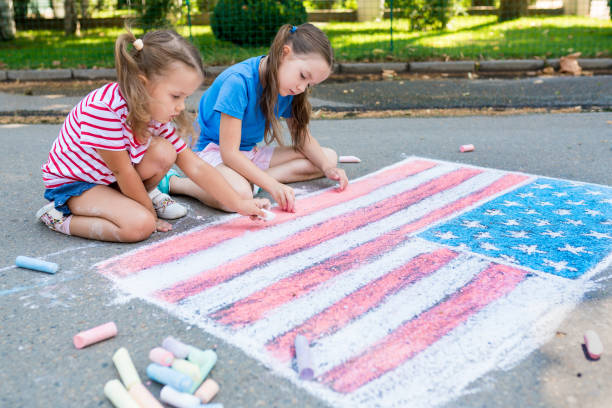 Two cute friends girls drawing American flag with colored chalks on the sidewalk near the house on sunny summer day. Kids painting outside. Creative development of children. Patriotic day concept Two cute friends girls drawing American flag with colored chalks on the sidewalk near the house on sunny summer day. Kids painting outside. Creative development of children. Patriotic day concept chalk drawing photos stock pictures, royalty-free photos & images