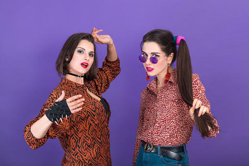 Two cute young women dressed in 80s style fooling around on violet background.