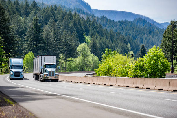 Two different big rig semi trucks with semi trailers running side by side on the winding multiline highway road with mountain and forest in Columbia Gorge stock photo