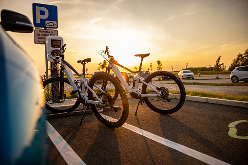 Two electric bicycles being charged at the electric vehicle charging station on parking lot during sunset