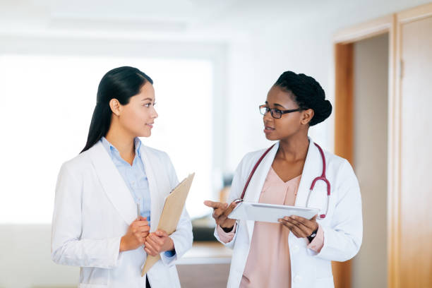 Two female doctors standing side by side and talking Two female doctors standing side by side in the hospital, holding a digital tablet and talking to each other. beautiful haitian women stock pictures, royalty-free photos & images