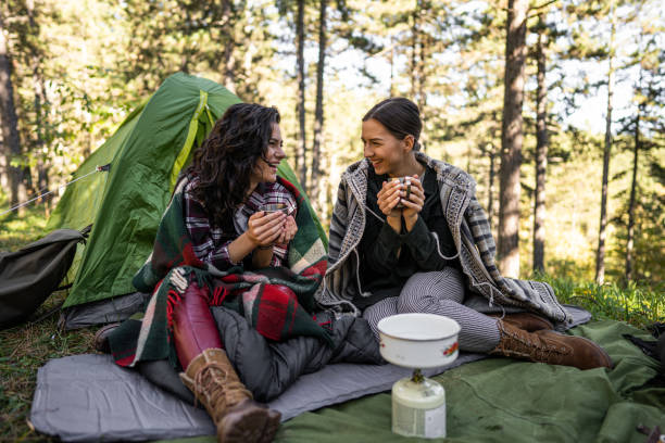 Two female friends sitting in front of tent in forest, smiling and drinking coffee stock photo