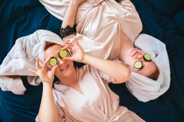 zwei weibliche models machen eine augenmaske mit frischgrüner gurke. zwei junge frauen in handtüchern und pyjamas haben eine lustige spa-party zusammen zu hause. - vladmodels stock-fotos und bilder