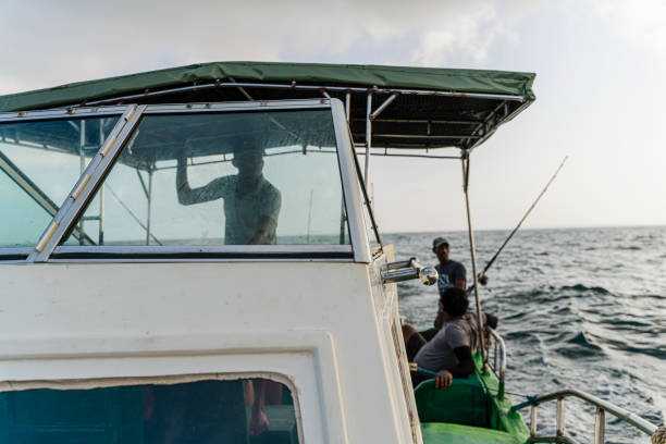 Two fishermen are resting and talking sitting at the stern of the fishing boat in the sea, waiting for a bite, when the captain is ruling the boat from a deckhouse. stock photo