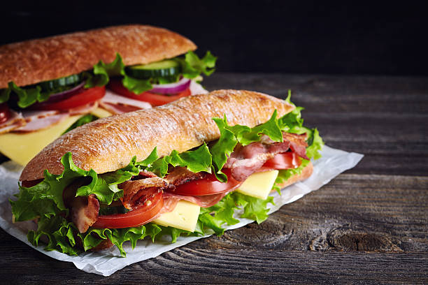 Two fresh submarine sandwiches Two fresh submarine sandwiches with ham, cheese, bacon, tomatoes, lettuce, cucumbers and onions on dark wooden background delicatessen stock pictures, royalty-free photos & images