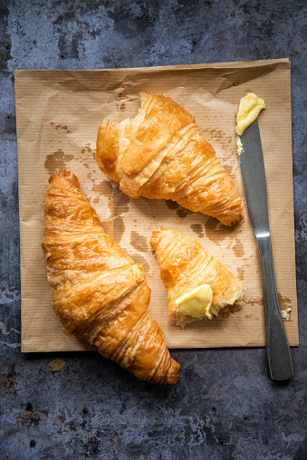 Two freshly baked French croissants are sitting on a brown paper bakers bag. One has been torn in half and some butter has been smothered across one end. There is a knife with butter on it resting on the bag beside the croissants. The background underneath them is a dark.