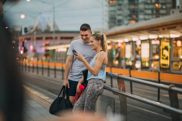 Two Friends Dressed in Athleisure Clothing Waiting for a Tram Two friends dressed in athleisure clothing waiting for a tram in Melbourne CBD, they are talking to one another and using a smartphone. federation square stock pictures, royalty-free photos & images