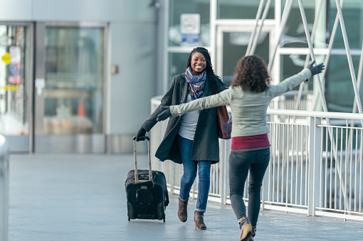 A pair of ethnic adult female friends are reuniting at at the airport and are happy. One of the women is rolling a suitcase while the other woman has her back to the camera and is holding her arms out for a hug.