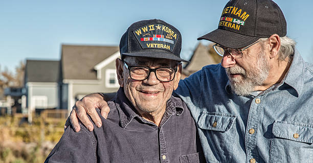 Two Generation Family USA Military War Veteran Senior Men Two generations of real person senior adult men USA military veterans. On the left, a 93 year old United States Army World War II and Korean Conflict US Air Force military veteran is smiling at the camera while his son-in-law on the right - a 65 year old US Navy sailor Vietnam War military veteran - is teasing him - trying to distract him by telling jokes during the photo shoot. They are both wearing generic souvenir shop military veteran commemorative baseball style caps. veteran stock pictures, royalty-free photos & images