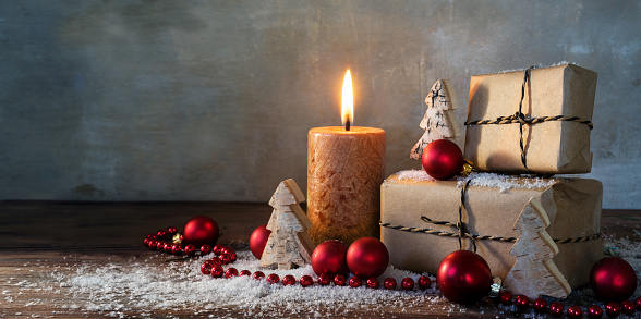 two gift boxes and a burning candle decorated with red christmas baubles and small wooden toy trees in some snow on rustic wood, vintage background with large copy space, panorama format, selected focus, narrow depth of field