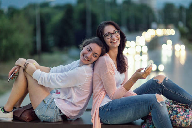 Two girlfriends sitting together with their phones in hands Two excited young women relaxing and leaning on each other backs, holding their mobile phones and laughing Portable DVD Player stock pictures, royalty-free photos & images