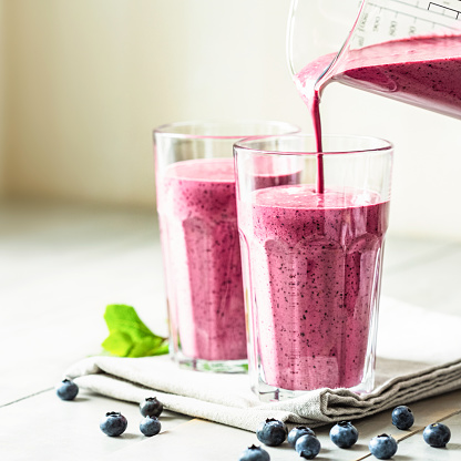 Two glasses of blueberry smoothie with mint garnish and straw on the table. Berry shake is poured into a glass.