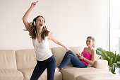 Adorable young woman in casual clothes wearing headphones singing song and funny dancing at home in front of smiling girlfriend sitting on sofa nearby with cellphone in hands. Teen girls enjoy music