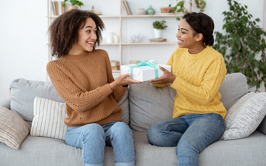Two ladies friends making surprise birthday present. Happy millennial african american female congratulates, gives gift to embarrassed mixed race sister, sits on sofa in interior of living room