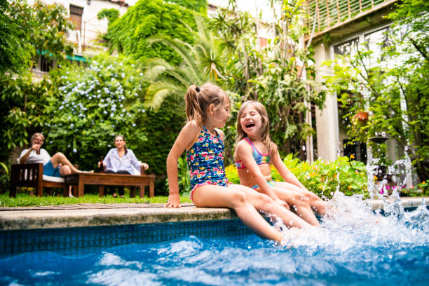 twee kleine meisjes zitten aan het zwembad en spetteren water met benen - zwembad stockfoto's en -beelden