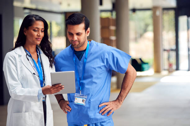 Two Medical Staff In White Coats And Scrubs With Digital Tablet Having Informal Meeting In Hospital stock photo