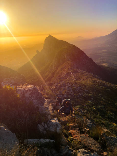 Two mid adult males hiking up a mountain ridge with rucksacks at sunset stock photo