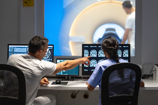 Rear view of male and female MRI technologist sitting at the console in the operating room and operating the MRI scanner