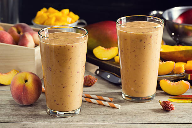 Two Peach Mango Smoothies in Glasses with Ingredients Two cold peach mango smoothies in pint glasses with striped straws. The peaches and mangoes are in the background in a colander and on a cutting board. Everything is on a faded wood picnic table. peach smoothie stock pictures, royalty-free photos & images