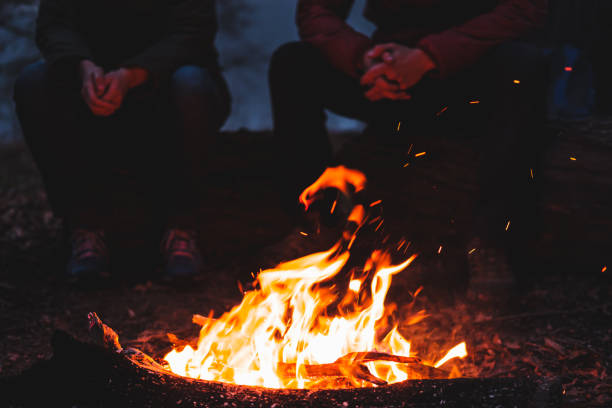 Two people sit by the bright bonfire at dusk. Spending nice time outdoors in chilly weather at a camping place - tranquil and peaceful scene Campfire stock pictures, royalty-free photos & images