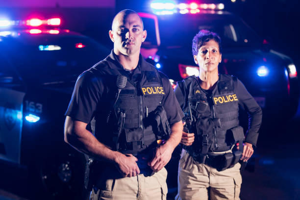 Two police officers standing in front of patrol cars Two multi-ethnic police officers standing in front of patrol cars, wearing bulletproof vests and duty belts. The policewoman is a mature African-American woman in her 40s. Her partner is a mid adult man in his 30s. It is nighttime and the emergency lights on top of the vehicles are flashing. armored clothing stock pictures, royalty-free photos & images