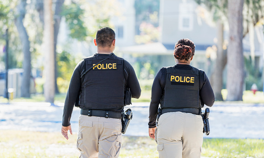 Community policing - rear view of two multi-ethnic police officers patrolling a local neighborhood on foot. They are walking side by side. The African-American policewoman is in her 40s. She and her partner, a young Hispanic man in his 20s.