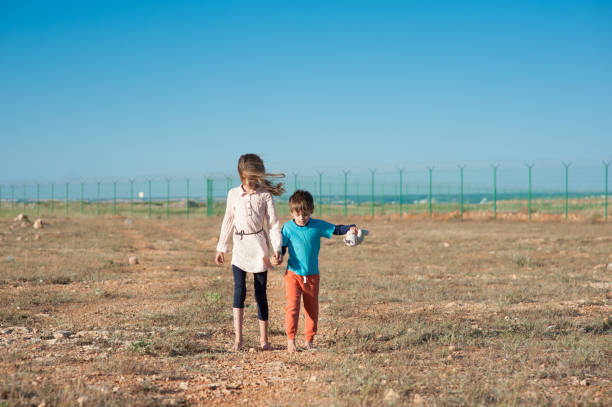 two poor refugee children seekers boy with toy and girl walking across hot desert with state border fence and blue sky on background hold hands two poor refugee children seekers boy with toy and girl walking across hot desert with state border fence and blue sky hand in hand hot mexican girls stock pictures, royalty-free photos & images