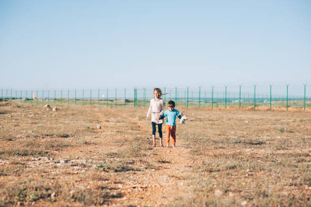 two poor refugee children seekers boy with toy and girl walking across hot desert with state border fence on background hand in hand two poor refugee children boy and girl walking across hot desert with state border fence on background hot mexican girls stock pictures, royalty-free photos & images