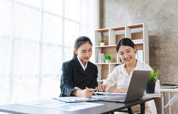 Two pretty young Asian businesswoman sitting at desk with laptop doing paperwork together discussing project financial report. Corporate business collaboration concept. Two pretty young Asian businesswoman sitting at desk with laptop doing paperwork together discussing project financial report. Corporate business collaboration concept. medical assistant online courses stock pictures, royalty-free photos & images