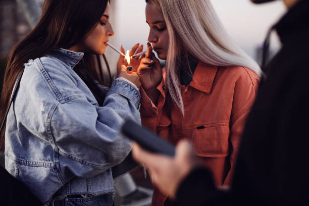 Two rebellious teenage girls are standing outdoors and lighting cigarettes. Teenagers smoking. stock photo
