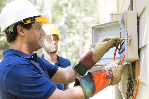 Caucasian and Latin men wearing hardhats and safety gloves work on the cable box connection for a residential location.  Younger worker sports a beard, they both wear uniform blue shirts.  Guy at box wears a safety face shield.