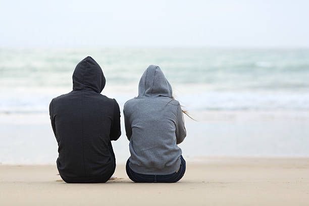 Two sad teenagers sitting on the beach Back view of two sad teenagers sitting on the sand of the beach and looking at horizon in a bad weather day depression land feature stock pictures, royalty-free photos & images