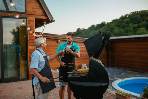 Two smiling men (young and old) drinking beer in the summer garden while barbecuing Senior man and son toasting with bottles of beer while barbecuing meat on the grill in the backyard fathers day stock pictures, royalty-free photos & images