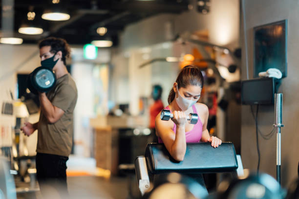 zwei sportler tragen schützende gesichtsmasken und trainieren im fitnessstudio, während sie die soziale entsagung aufrecht erhalten - fitnesseinrichtung stock-fotos und bilder