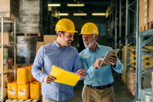 Two successful smiling business man walking through big warehouse with helmets on their heads. Older man is holding digital tablet and shoving younger one some documents. Two successful smiling business man walking through big warehouse with helmets on their heads. Older man is holding digital tablet and shoving younger one some documents. two generation family stock pictures, royalty-free photos & images