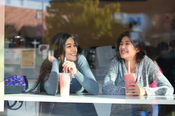 Two teen girls at outdoor cafe drinking boba tea together stock photo