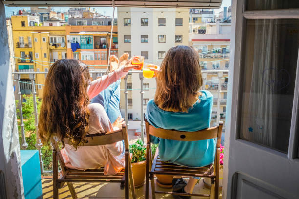 Two teenage girls, toasting with natural juices and sunbathing , sitting on chairs on the balcony , enjoying the cityscape stock photo